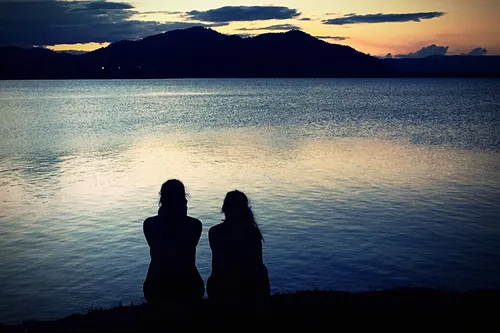 photo of two woman sitting by lake at sunset
